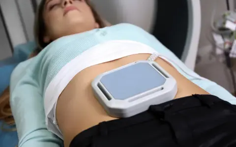 Kobieta leży podczas zabiegu, na brzuchu ma nałożone urządzenie do terapii polem magnetycznym, co jest częścią jej rehabilitacji.