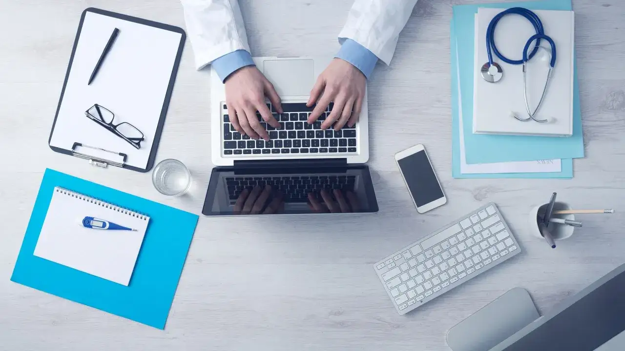 Lekarz pisze na laptopie, obok stetoskop, telefon i notatnik z termometrem. Ważne skierowanie do specjalisty to klucz do szybkiej diagnozy.